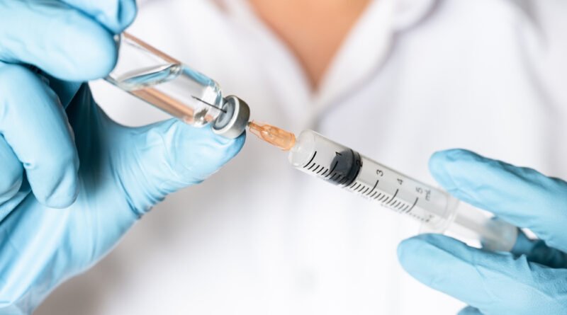 Mano con guante sosteniendo una jeringa y un vial, ilustra la campaña de vacuna antigripal